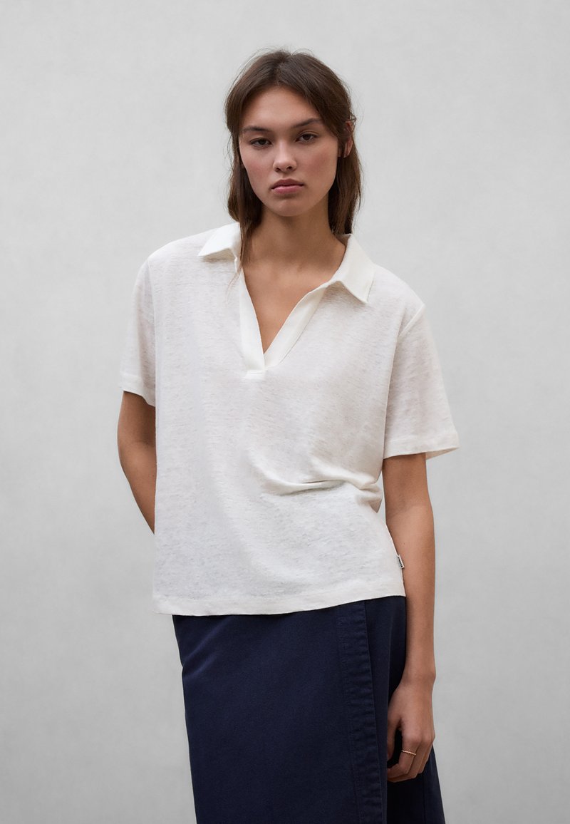 Jauna sieviete ar vidēja garuma brūniem matiem, valkājot vaļīgu baltu īsroku krekliņu ar apkakli un tumšu svārku pret pelēku fonu.