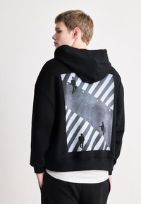 Sudadera negra con una gran impresión gráfica en la parte posterior, que representa figuras en un paso de cebra a rayas. El texto "HUGO" aparece dentro del diseño.