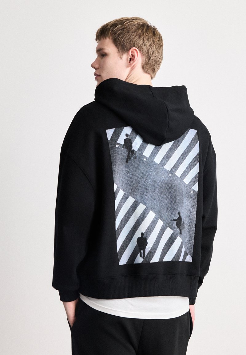 Sudadera negra con una gran impresión gráfica en la parte posterior, que representa figuras en un paso de cebra a rayas. El texto "HUGO" aparece dentro del diseño.
