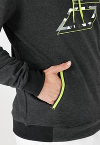 Sudadera gris oscuro con un bolsillo resaltado por una cremallera verde lima. Cuenta con puños acanalados y un logo geométrico en blanco y gris.