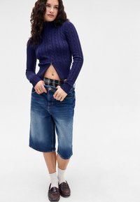 Purple cable-knit cropped sweater with a high neck, paired with light blue denim shorts ending below the knee and brown loafers.