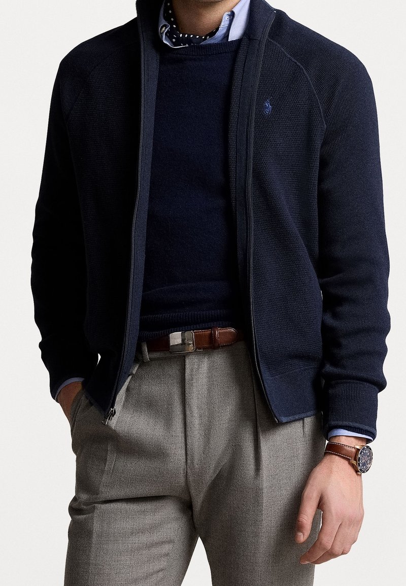 Marineblauwe sweater met rits en een gestructureerde afwerking, gedragen over een donkerblauw shirt met ronde hals. Grijze broek met een bruine riem en horloge, gecombineerd met een nette overhemdkraag.