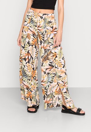 Femme portant un pantalon à jambes larges et taille haute avec un imprimé de feuilles tropicales, associé à des sandales noires et un haut court noir, posant devant un fond blanc.