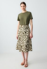 T-shirt verde oliva abbinata a una gonna beige con una stampa astratta in olive, sandali flat e gioielli semplici.