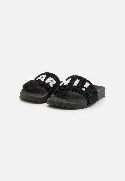Sandalias de slide negras con tiras de tela texturizada que presentan letras blancas. Suelas de goma sólidas que brindan agarre y comodidad. Diseño casual adecuado para uso en interiores y exteriores.