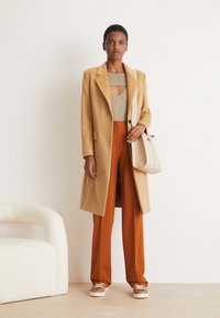 Cappotto di lana beige sopra un top grigio a maglia e pantaloni arancioni a gamba larga. Gli accessori includono una borsa beige chiaro e sneakers marroni con lacci.
