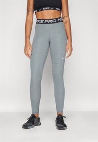 Legginsy Nike Pro w kolorze heather gray, dopasowane z płaskimi szwami. Zawierają czarny pas z powtarzającym się tekstem "Nike Pro" i małym logo.