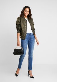 Sieviete, kura valkā olīvu krāsas bomber jakas, baltu kreklu, zilo skinny džinsu, melnas kurpes ar augstu papēdi un tur rokās melnu rokassomu ar zelta ķēdes siksniņu.