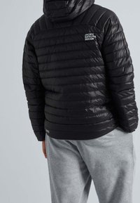 Veste matelassée noire avec capuche, présentant une texture matelassée et un logo sur le haut du dos. Associée à un pantalon de survêtement gris.