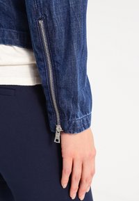 Gros plan sur le bras d'une personne portant une veste en jean bleue avec une fermeture éclair argentée sur la manche et un pantalon bleu foncé.