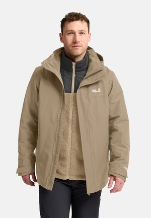 Man draagt een beige buitenjas over een lichtbruine fleece met zwarte kraag, staand tegen een effen lichte achtergrond, kijkt recht vooruit.