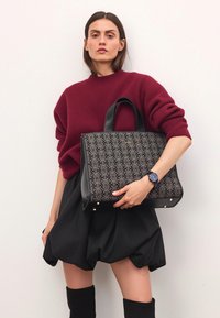 Sort mønstret tote bag med læderdetaljer, båret af en person iført en bordeaux sweater og en sort nederdel, mod en neutral baggrund.