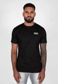 Sort bomuld t-shirt med korte ærmer og rund udskæring, med et hvidt logo på venstre bryst. Enkelt design, tætsiddende silhuet.