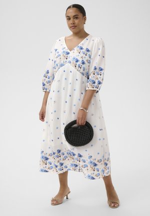 Femme portant une robe midi blanche avec des motifs floraux bleus, des sandales à talons beige, tenant un sac à main rond en osier noir à ses côtés.