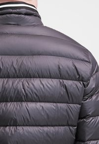 Chaqueta negra tipo plumífero con diseño acolchado, que presenta costuras horizontales, un cuello alto y un detalle de punto a rayas en el cuello.