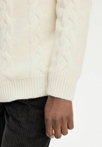 Maglione a trecce in bianco sporco, con pomelli alti e orlo a costine. Abbinato a pantaloni in velluto a coste neri con trama.