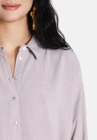 Camicia button-up di un leggero colore viola con un tessuto texturizzato, caratterizzata da un colletto e bottoni a contrasto. Orecchino giallo visibile sul lato sinistro.