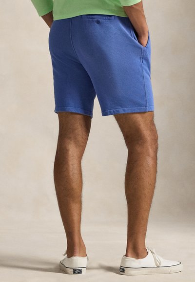 Homme portant un short bleu, un t-shirt vert et des baskets blanches, vu de dos les mains dans les poches sur un fond neutre.