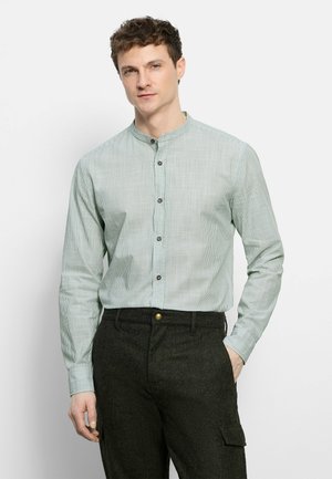 Homme portant une chemise rayée vert clair à col mandarin et un pantalon foncé, debout avec une main dans la poche devant un fond uni.