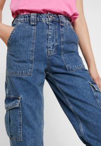 Pantalones cargo de mezclilla azul con cintura alta, cintura elástica y múltiples bolsillos grandes. La tela tiene textura con un aspecto clásico desgastado.