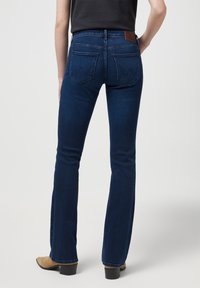 Jean évasé bleu foncé avec une taille haute, présentant des poches arrière et une étiquette de marque en cuir brun. Texture en denim lisse.