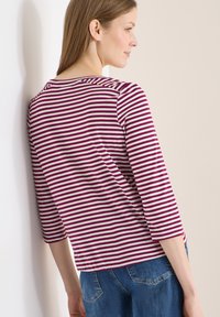 Femme portant un chemisier à manches longues rayé violet et blanc et un jean bleu, appuyée contre un mur clair, de profil.