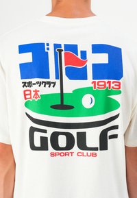 Valkoinen t-paita, jossa on monivärinen graafinen printti golf-teemalla, mukaan lukien vihreä golfkenttä, lippu ja tekstielementit sinisessä ja punaisessa.