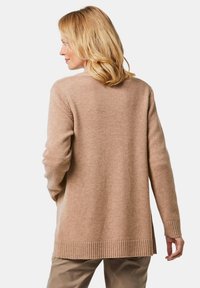 Een lichtbruine cardigan met geribbelde manchetten en zoom, met een relaxte pasvorm en een gestructureerde breisel. Zichtbare zijsplitten verbeteren het comfort en de stijl.