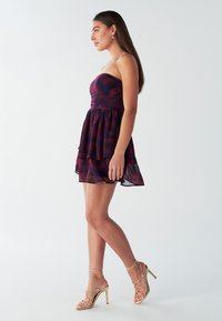 Strapless jurk in een maroon en marineblauw camouflagepatroon, met een getailleerde lijfje en gelaagde, semi-transparante rok. Gouden strap heels.