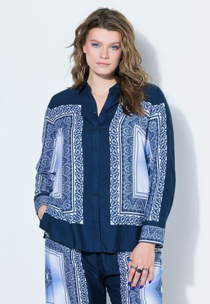 Young woman with wavy hair wears matching blue and white patterned shirt and pants, standing with one hand in pocket against white wall.