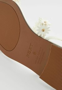 Weiße Sandalensohle mit floralen Verzierungen, goldenen Akzenten und einer strukturierten braunen Gummisohle; versehen mit dem „NEXT“-Logo und Größenangaben.