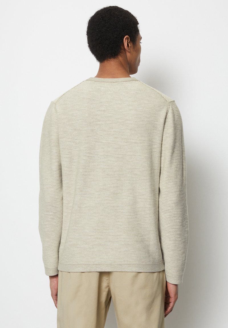 Beżowy sweter o fakturze z długimi rękawami, okrągłym dekoltem i prążkowanymi detalami na ramionach, zestawiony z jasnobeżowymi spodniami.