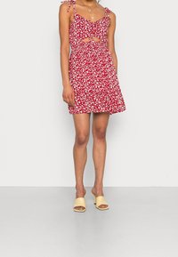 Röd blommig klänning med tunna axelband, utskärning vid midjan och en utsvängd kjol; matchad med gula klackmules.