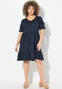 Tummansininen puuvillamekko lyhyillä hihoilla, V-kaula-aukolla ja kerroksilla varustetulla röyhelöhelmalla. Yhdistetty vaaleanruskeisiin korkokenkiin. Sileä pinta, rento muotoilu.