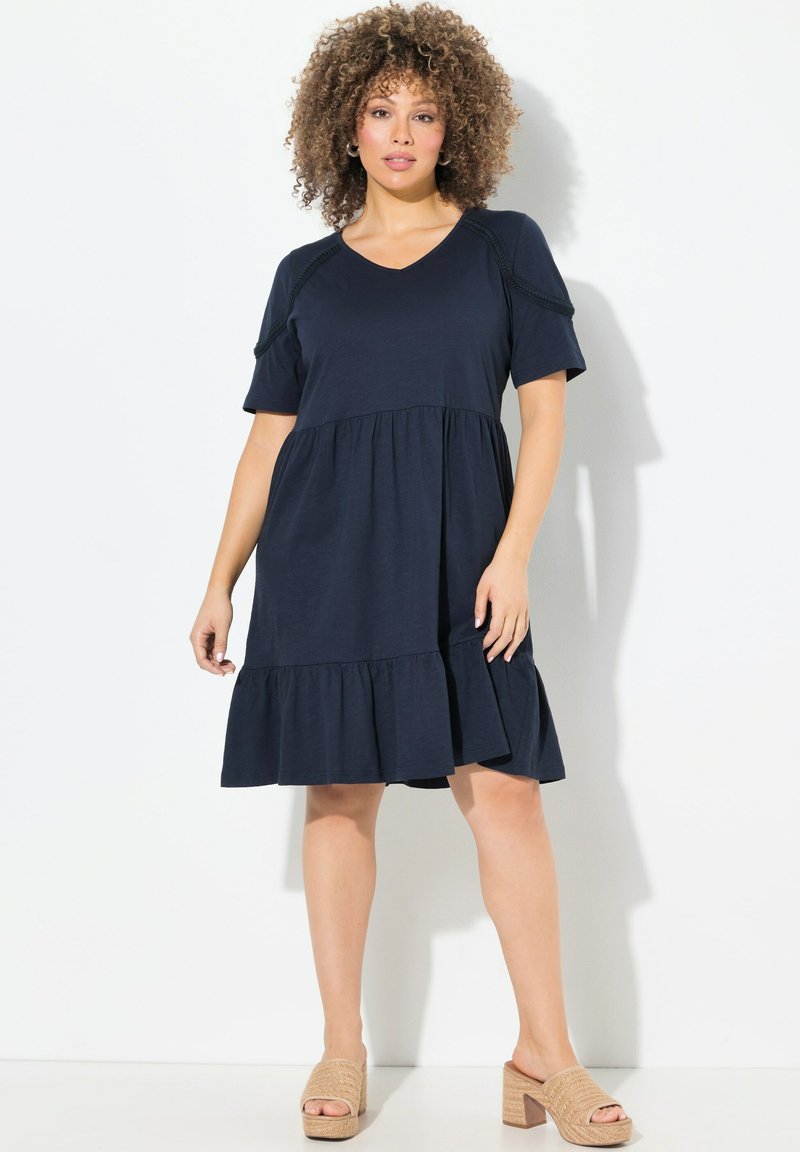 Tummansininen puuvillamekko lyhyillä hihoilla, V-kaula-aukolla ja kerroksilla varustetulla röyhelöhelmalla. Yhdistetty vaaleanruskeisiin korkokenkiin. Sileä pinta, rento muotoilu.