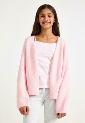 Dívka se usmívá, má na sobě jemný růžový oversize cardigan přes světle růžový pruhovaný top a světle šedé kalhoty se stahovací šňůrkou, stojící před jednobarevným pozadím.