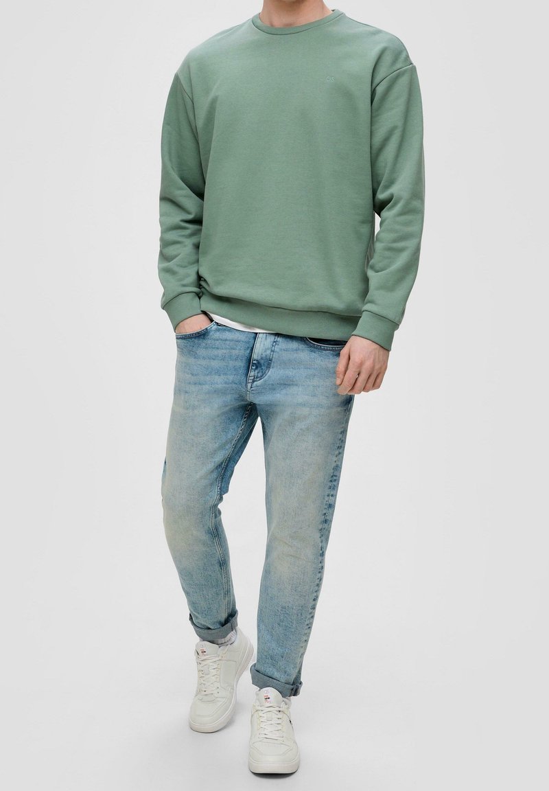 Sudadera verde con cuello redondo, de tejido ligero; jeans de mezclilla azul claro, de corte ajustado; zapatillas blancas con detalles texturizados.