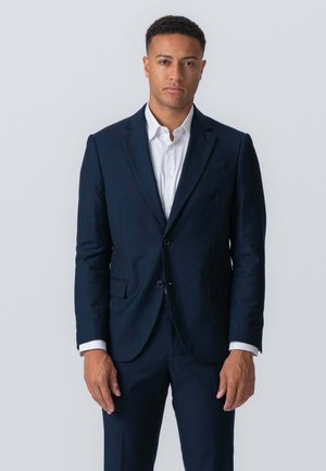 Marineblå jakkesæt med to-knaps design og klapkrave, parret med en hvid skjorte. Glat tekstur, skræddersyet pasform og minimalistisk stil.