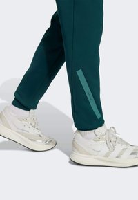 Pantalones de chándal en un verde azulado oscuro con un corte ajustado que cuentan con una franja lateral de acento. Combinados con zapatillas deportivas blancas con detalles plateados y cordones.