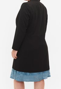 Veste noire à manches longues portée sur une robe en dentelle bleu clair, présentant un ourlet en dentelle transparent, une texture lisse et une coupe ajustée.