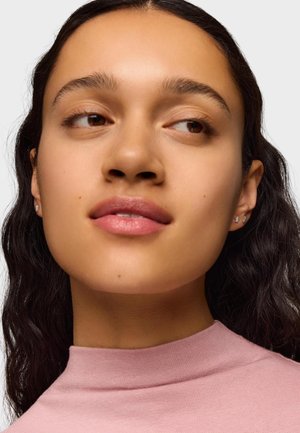 Retrato de una persona con cabello oscuro y ondulado, que lleva una blusa rosa claro con cuello alto. Un maquillaje sutil destaca los labios y las mejillas. Pendientes de aro de plata.