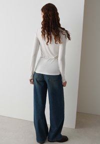 Langärmliges weißes Oberteil, kombiniert mit lockeren, dunkelblauen Denim-Jeans. Der Stoff wirkt weich und glatt, mit einer entspannten Silhouette.