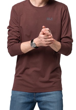 Osoba ubrana w brązową bluzkę z długim rękawem Jack Wolfskin i niebieskie dżinsy, trzymająca złożone dłonie z przodu, z czarnym zegarkiem na lewym nadgarstku.