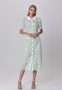 Robe en dentelle vert menthe avec des manches courtes bouffantes, un col Peter Pan blanc, un devant boutonné et un motif floral, terminée par un ourlet à volants.
