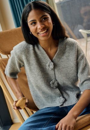 Grau gestrickte Strickjacke mit blumenförmigen Knöpfen, kurzen Ärmeln, geripptem Saum, getragen über blauen Jeans. Weiche Textur und lässige Passform.