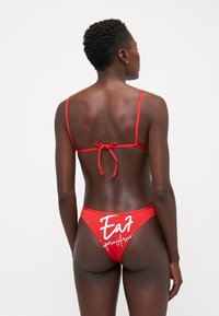 Crveni bikini set s tankim, prilagodljivim trakama i zatvaranjem na vezanje. Donji dio ima natpise "Eat" i "Superior Aqua" u bijeloj boji.