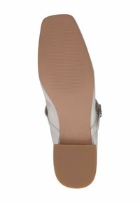 Suela de zapato plana con una superficie de goma texturizada de color marrón claro, que presenta una correa en el tobillo y un material superior suave de color blanco roto.