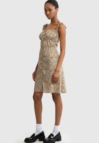 Blumenkleid mit verstellbaren Trägern, ausgestattet mit einem gerafften Oberteil und einem gestuften Rock in Brauntönen, Orange und Weiß. In Kombination mit schwarzen Schuhen.