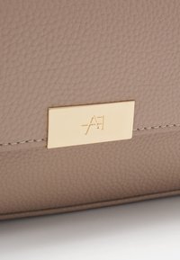 Beżowa torba skórzana o teksturowanej powierzchni, z metalową złotą płytką z logo. Proste przeszycia wzdłuż brzegów podkreślają design.