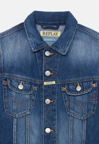 Chaqueta de mezclilla en azul oscuro con un cuello clásico, botones plateados y dos bolsillos en el pecho con detalles de costura. Etiqueta "REPLAY" en el cuello.